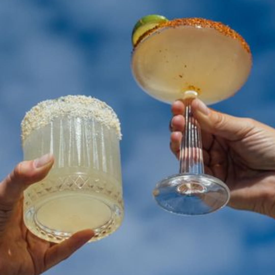 Two hands holding margaritas against a blue sky.