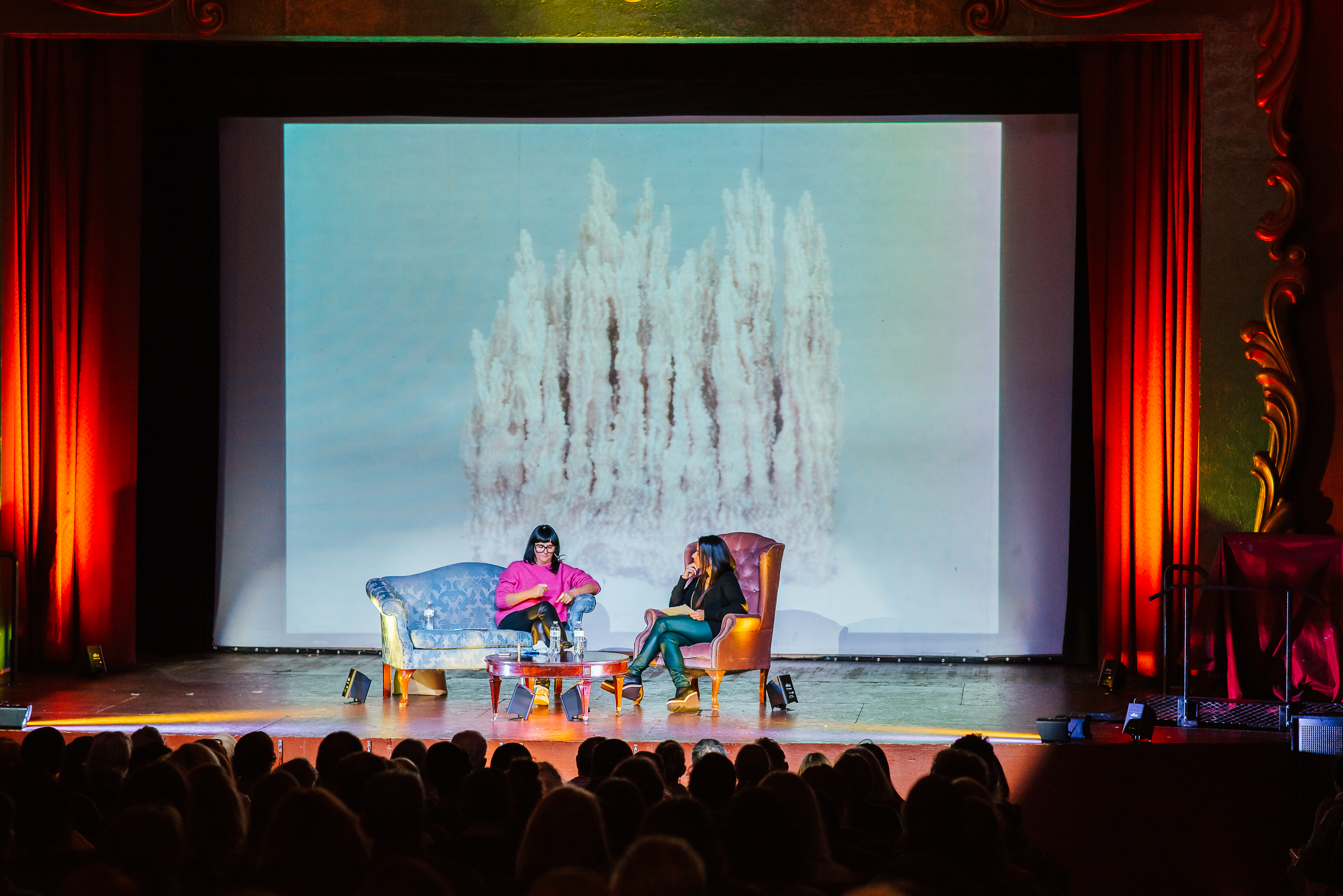 Stage in a performance space with two women sitting on vintage chairs in front of a sculptural work displayed on large screen tucked between red floor length curtains. The audience members are facing the stage, and only the stage is lit up. 