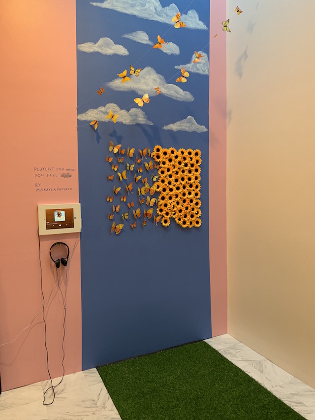 Green turf is laid out on the floor. The wall is painted blue and sunflowers are glued to it. The sunflowers morph into yellow butterflies and fly off into the sky. 