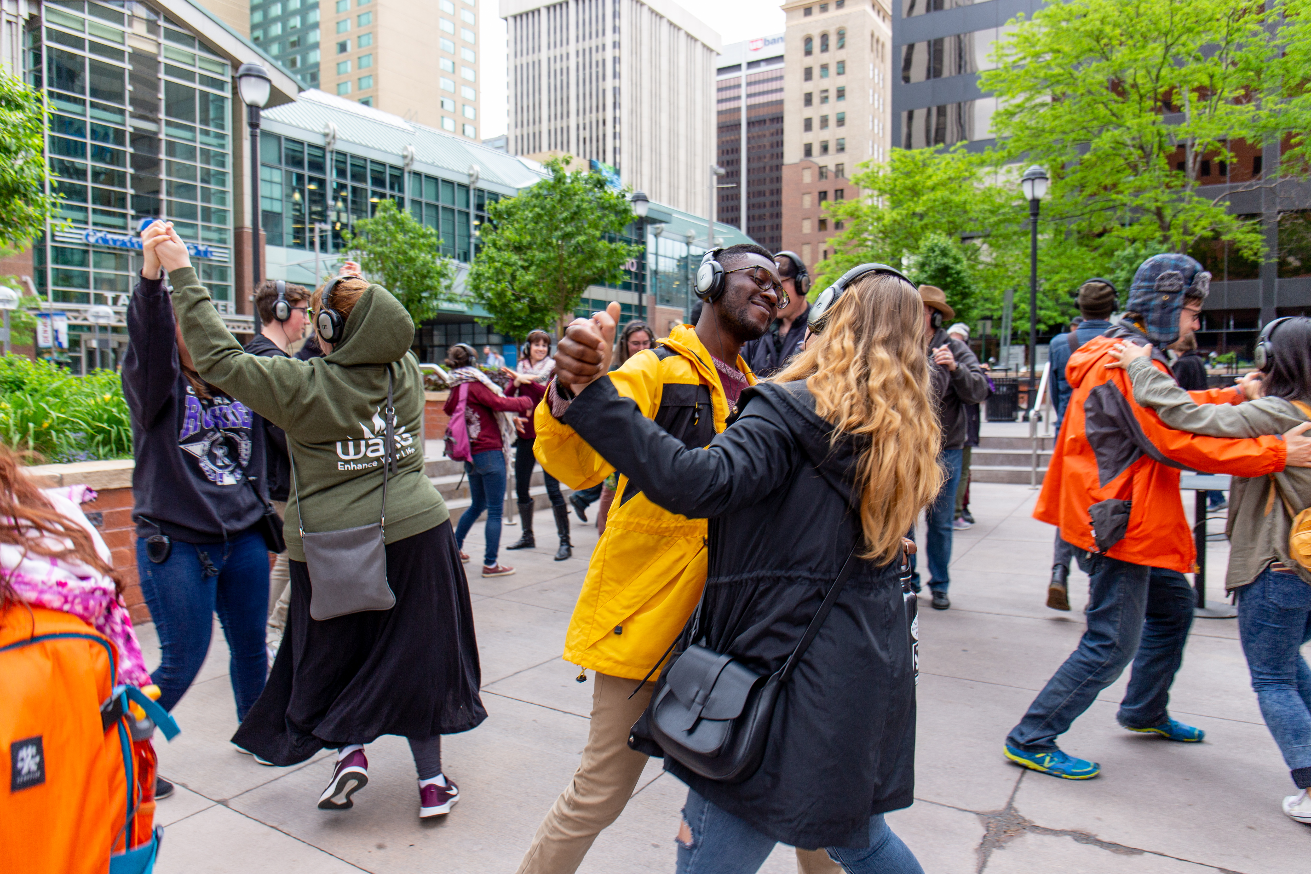 A diverse group of people wear headphones and dance with each other in an urban area. 