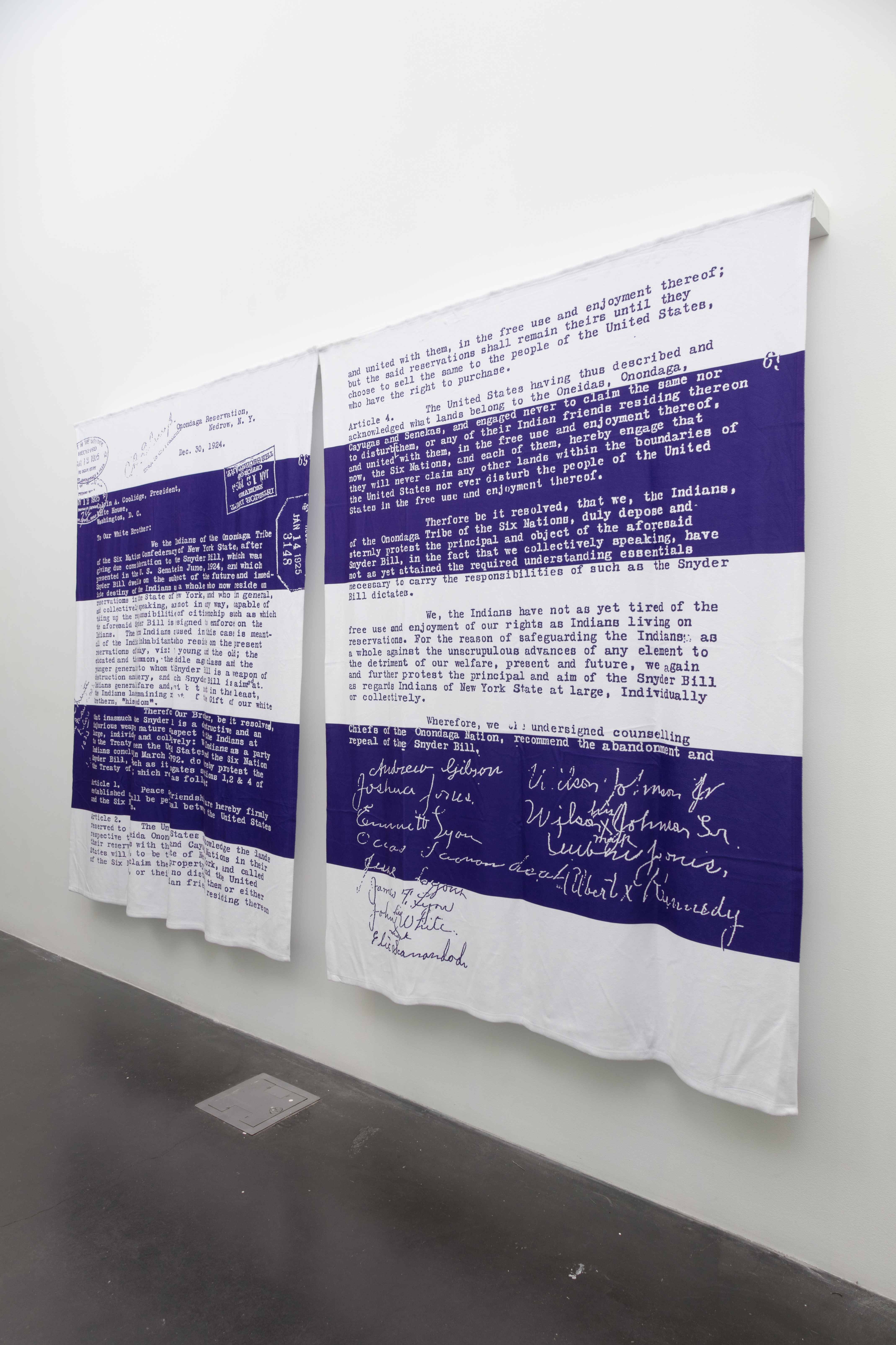  [Image description Artist Alan Michelson's, artwork of a reproduction of the December 30, 1924 letter to Calvin Coolidge by the chiefs of the Onondaga Nation, protesting the Indian Citizenship Act and reaffirming Onondaga sovereignty. The reproduction is on a pair of blankets in the colors white and purple and hangs on a white gallery wall with a dark concrete floor.]