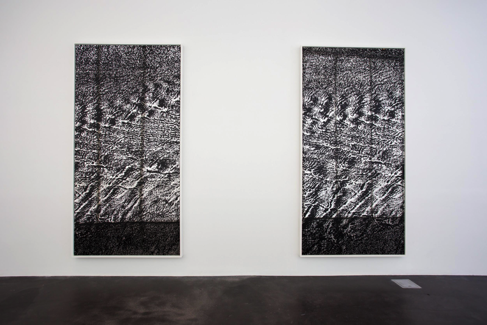 Two abstract grainy artworks in black and white resemble sand dunes are waves of water. They hang vertically against a white wall in a gallery.
