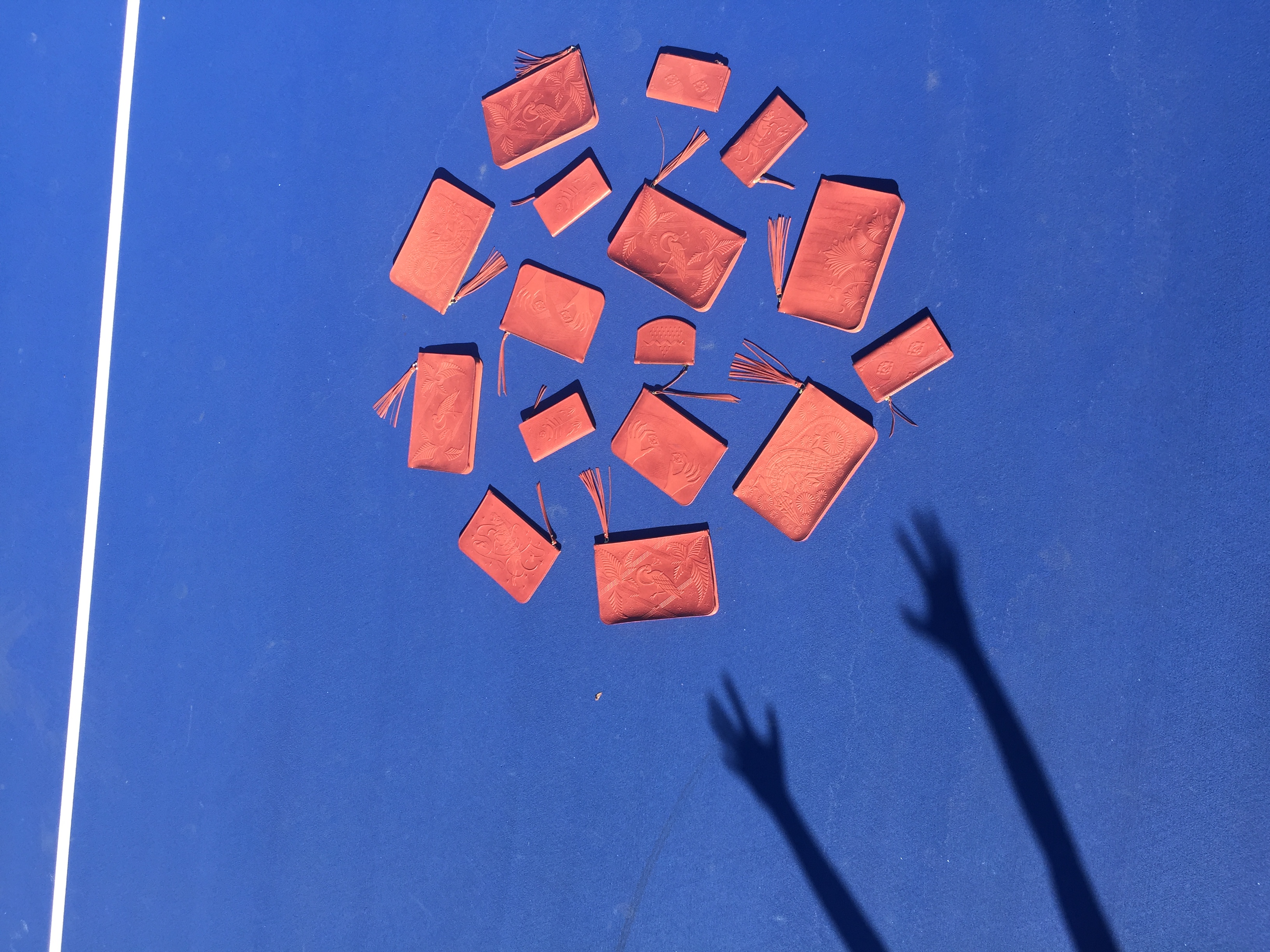 Several red wallets and clutches placed in a circle and photographed on a blue colored floor which looks like it could be a tennis court. On the left side of the frame is a shadow of two arms, which look like they tossed the red wallets and clutches on the tennis court. 