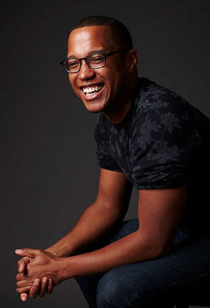 Playwright, Branden Jacobs Jenkins, sitting angled in blue jeans and dark tee shirt, hands clasped and his has a joyful smile. 