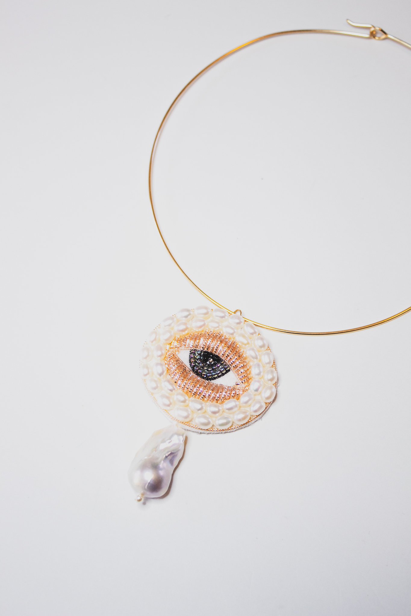 Necklace photographed on a stark white background. The necklace features an embroidered eye, using metals used to embellish the surface. There are pearl-like obejcts surrounded the eye and a larger stone hanging from it. 