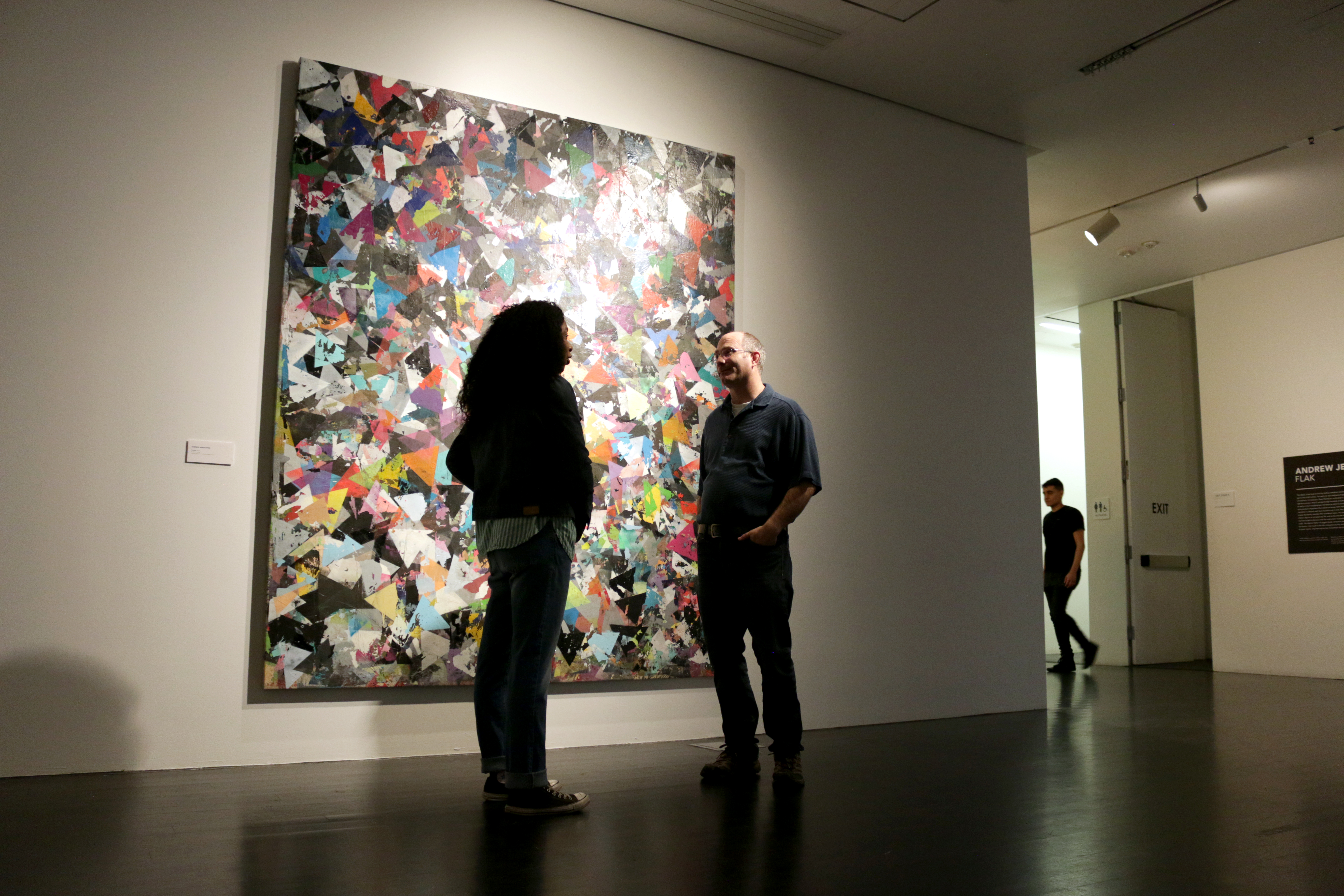 Two people talk ﻿in front of an abstract artwork in a dimly lit gallery. 