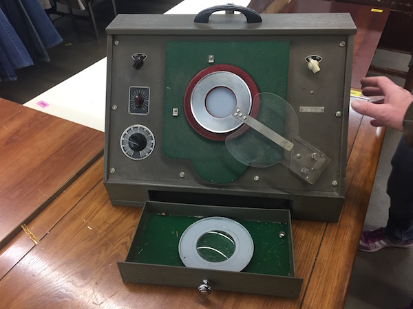 An old electronic instrument of unknown use or purpose. It has three dials on the side and a lens in the center. It is dark green with a handle on the top. 