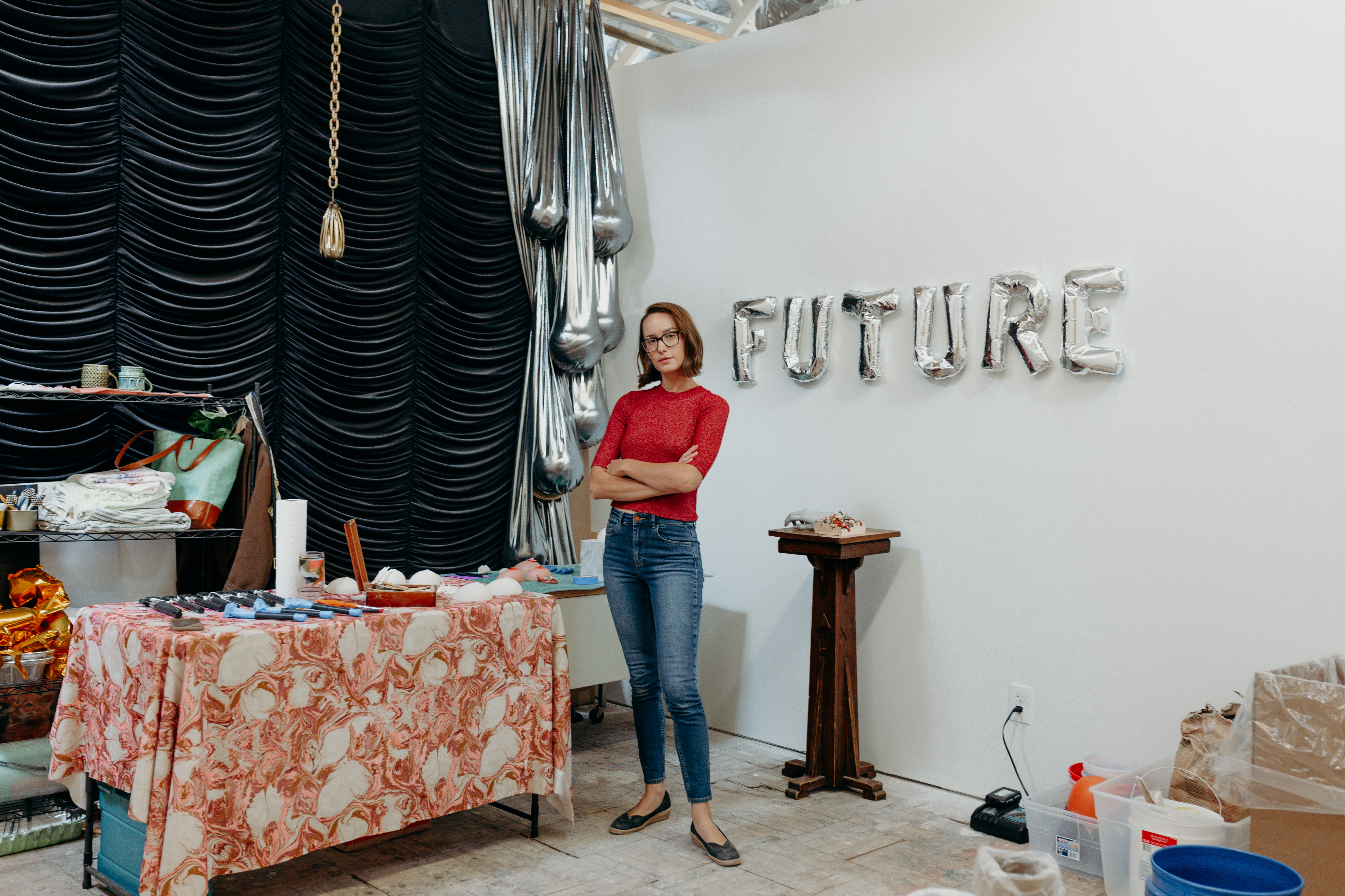Laura Shills poses in her studio with her arms crossed in front of her. Behind her on a white wall are silver balloons spelling out the word Future. 
