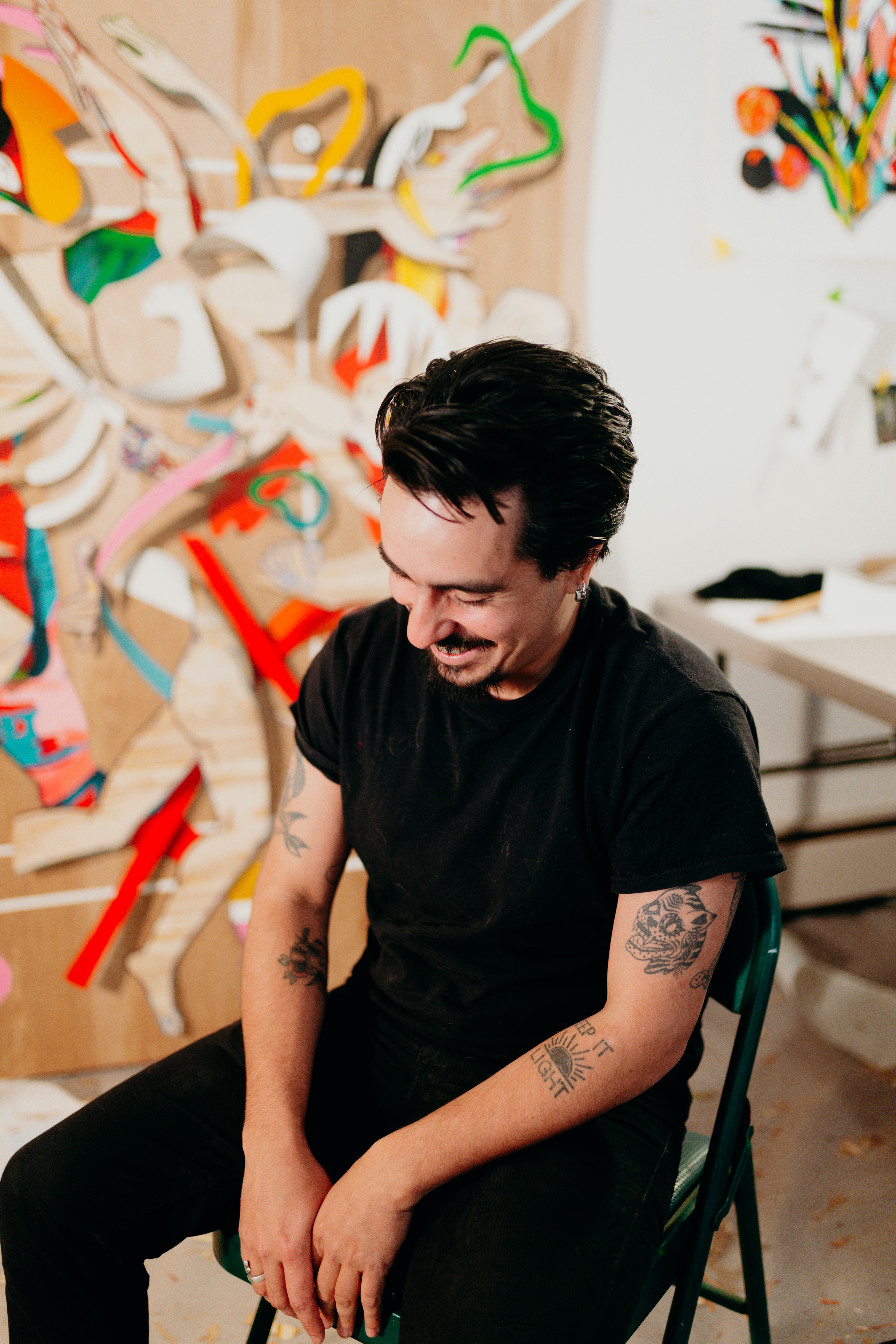 Portrait of a man sitting on a chair in a studio space. He is wearing a black t shirt and black pants. He is mid laugh