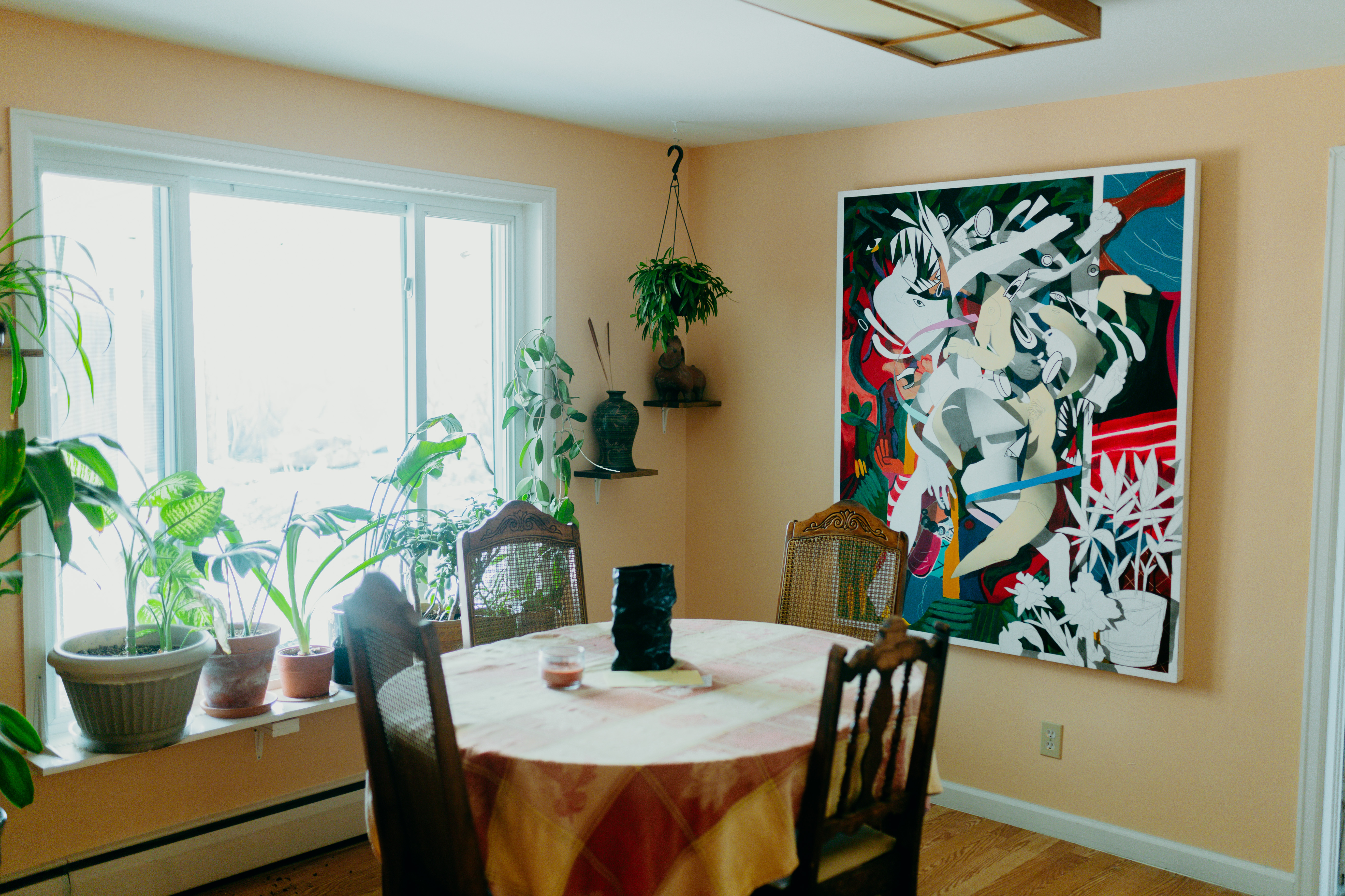 A dining room filled with plants. On the wall is a large abstract painting. A round table with four chairs rests in the center of the room. 