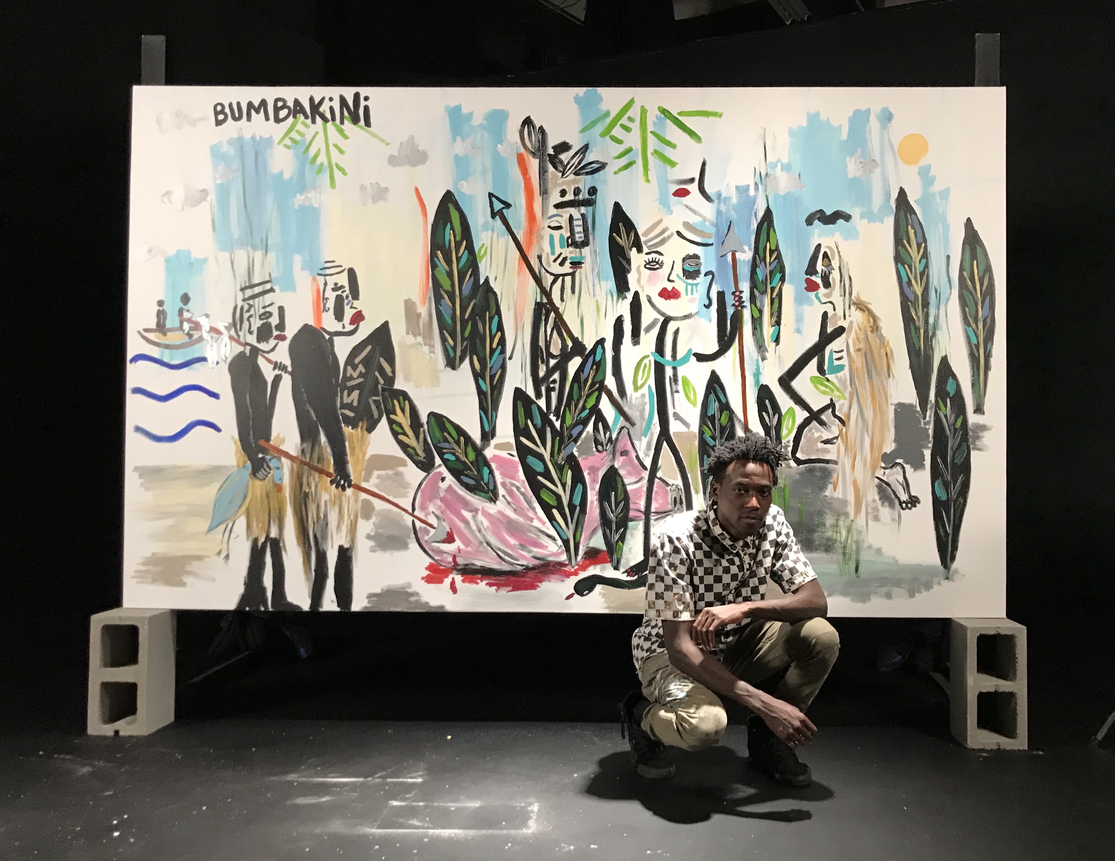 A photo of artist Lio crouched in front of one of his large colorful works on canvas, which is sitting on two concrete cinderblocks. The painting on the canvas is inspired by the Congo and the people there. 