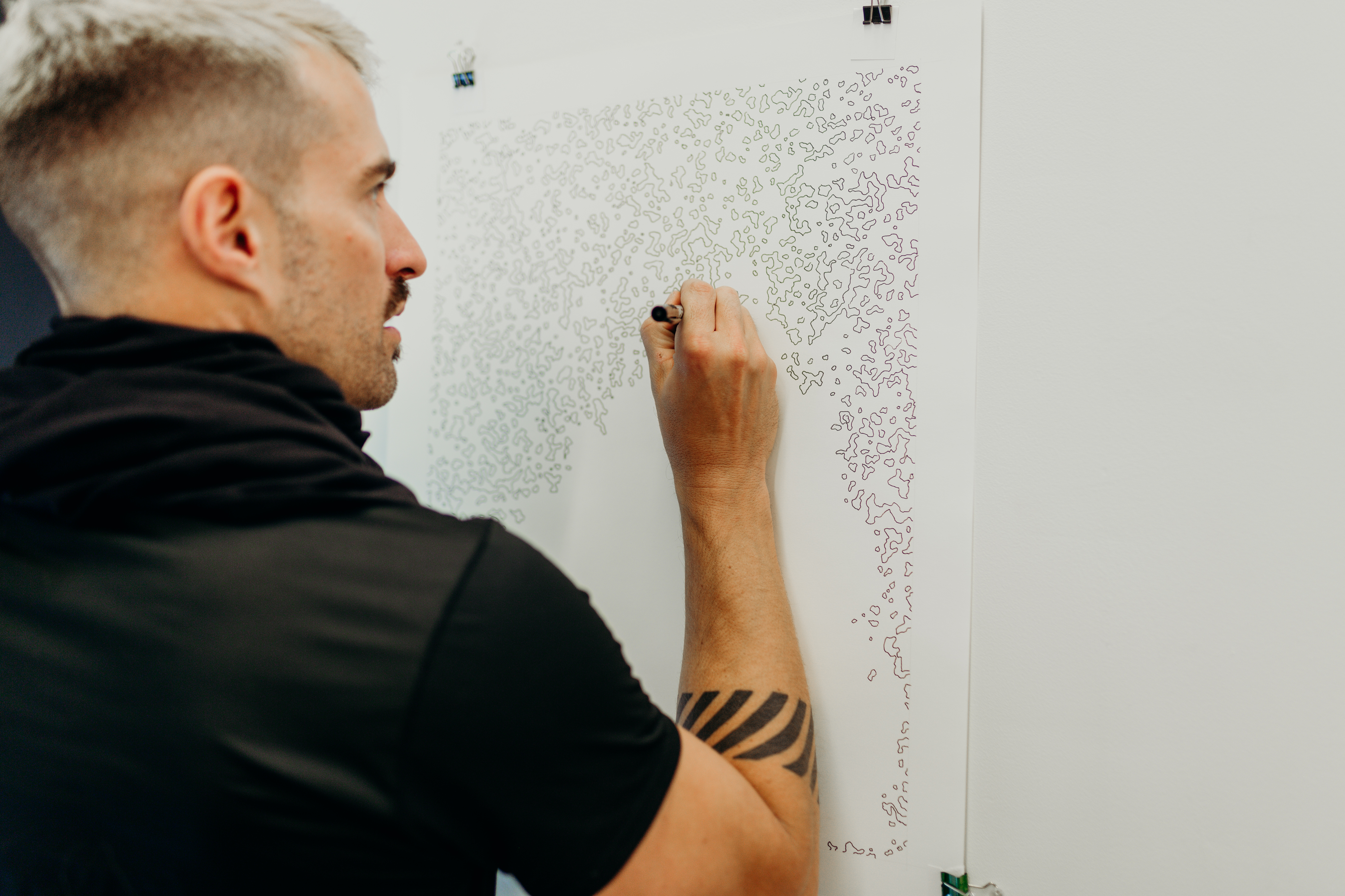 Candid shot of joel swanson drawing on a piece of paper hung on the wall. The pattern is abstract composed of several small wobbly shapes. 