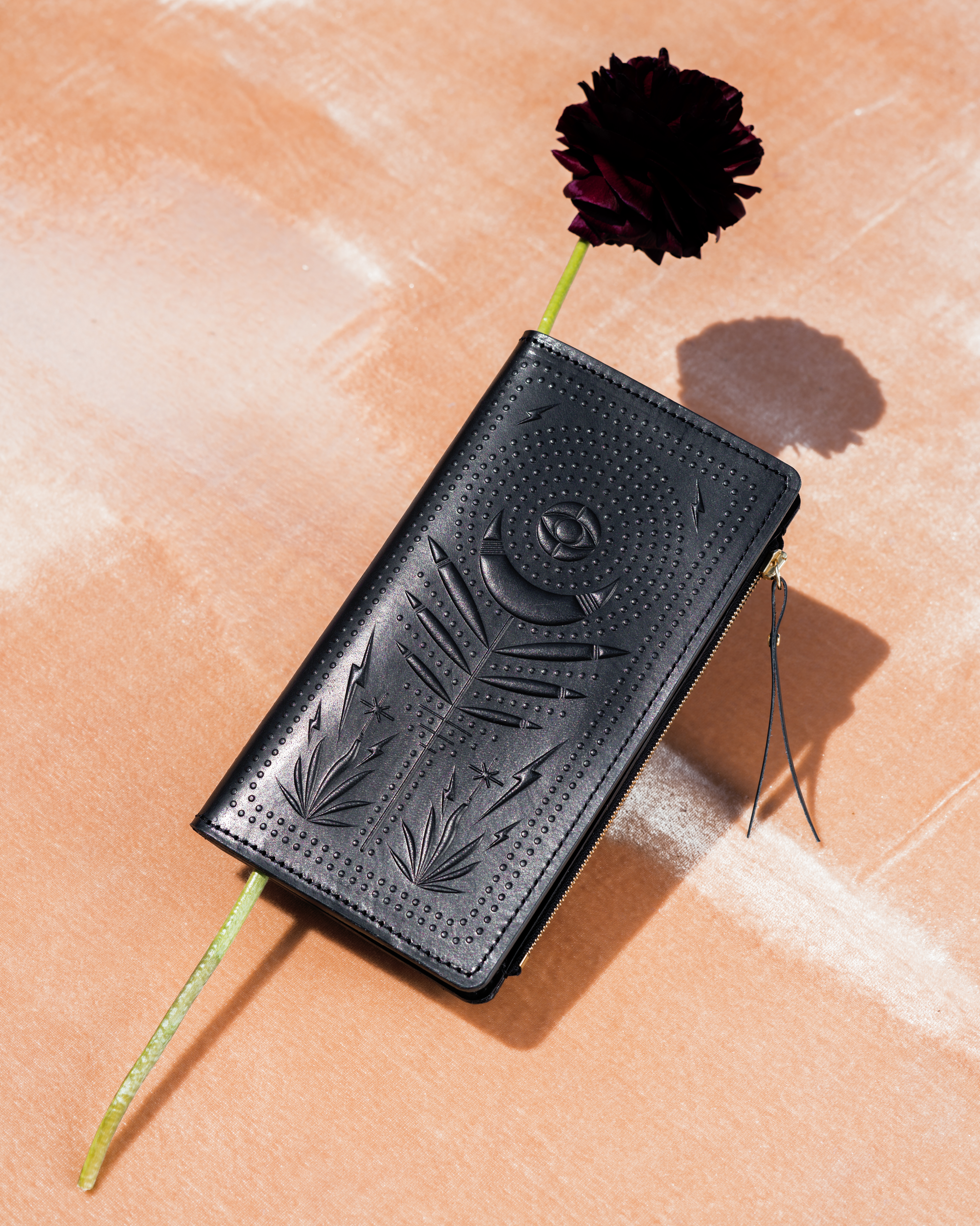 Black zip wallet with mystical design and a flower tucked inside the wallet. It’s photographed under harsh light, which casts a shadow on the pink velvet backdrop the wallet is photographed on. 