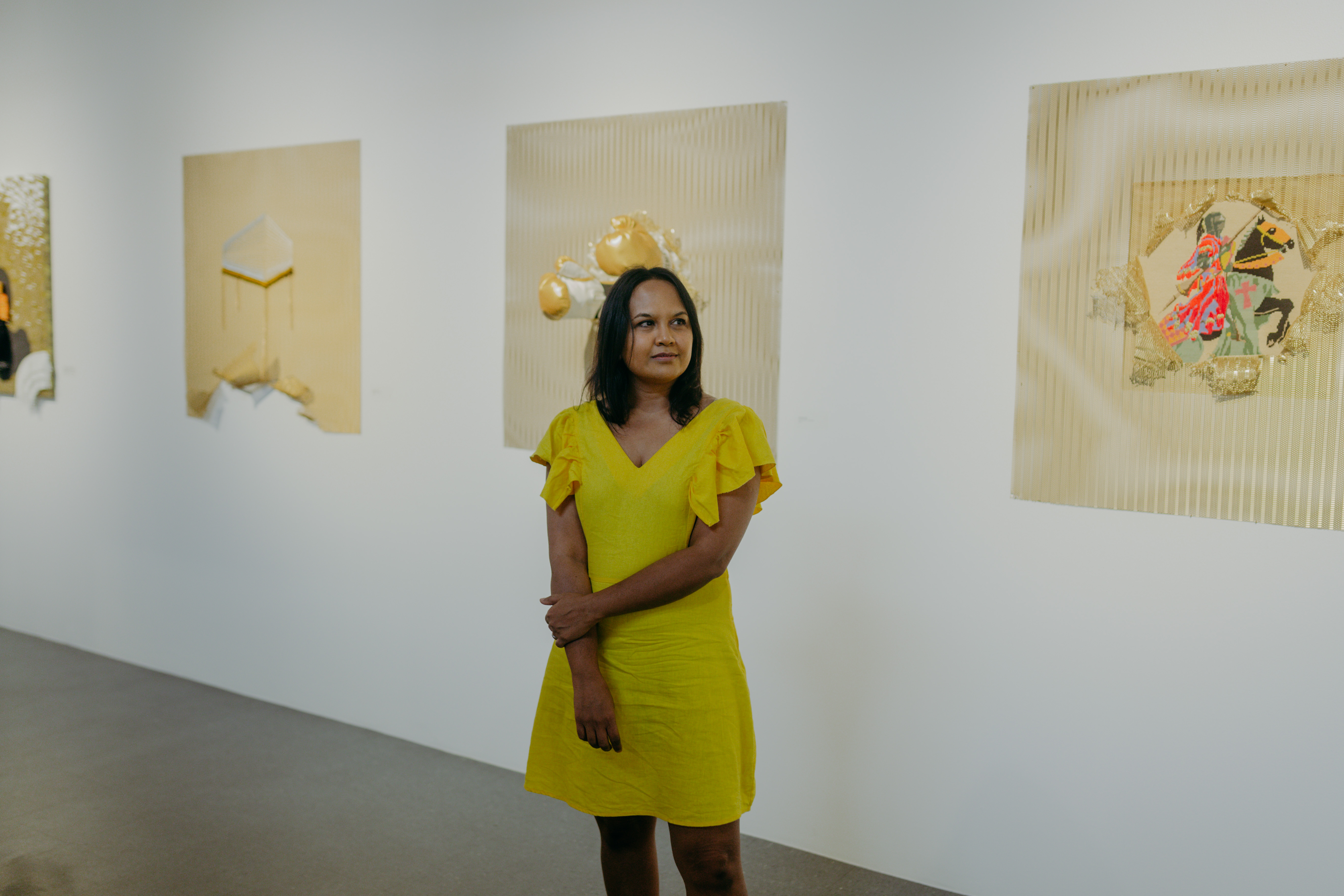 Sucitra stands in front of artworks wearing a bright yellow dress. ﻿