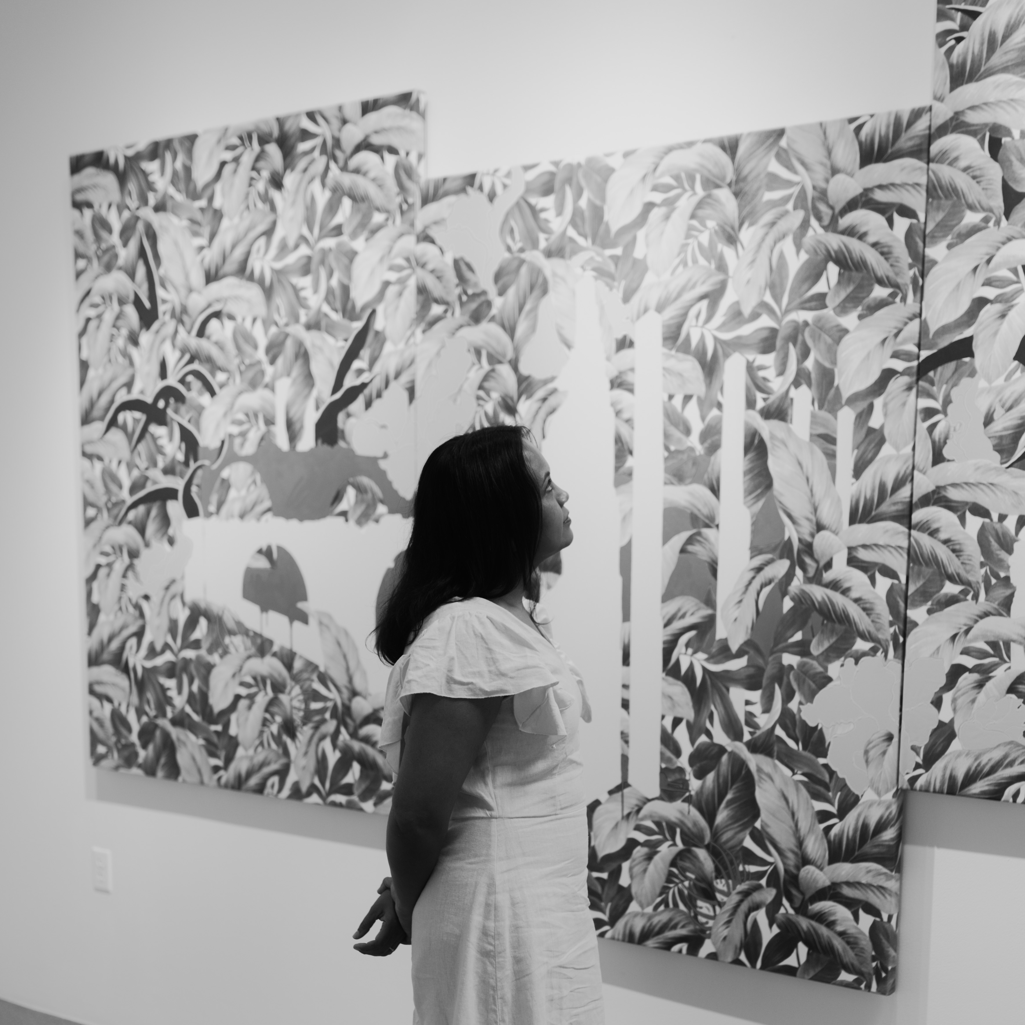 Black and white portrait of Suchitra looking at her three paneled artwork of fern leaves. 