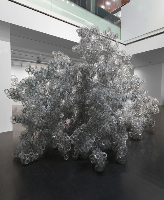 A large amorphous sculpture made up entirely of slinkies rests in a gallery space upon dark wood flooring. 