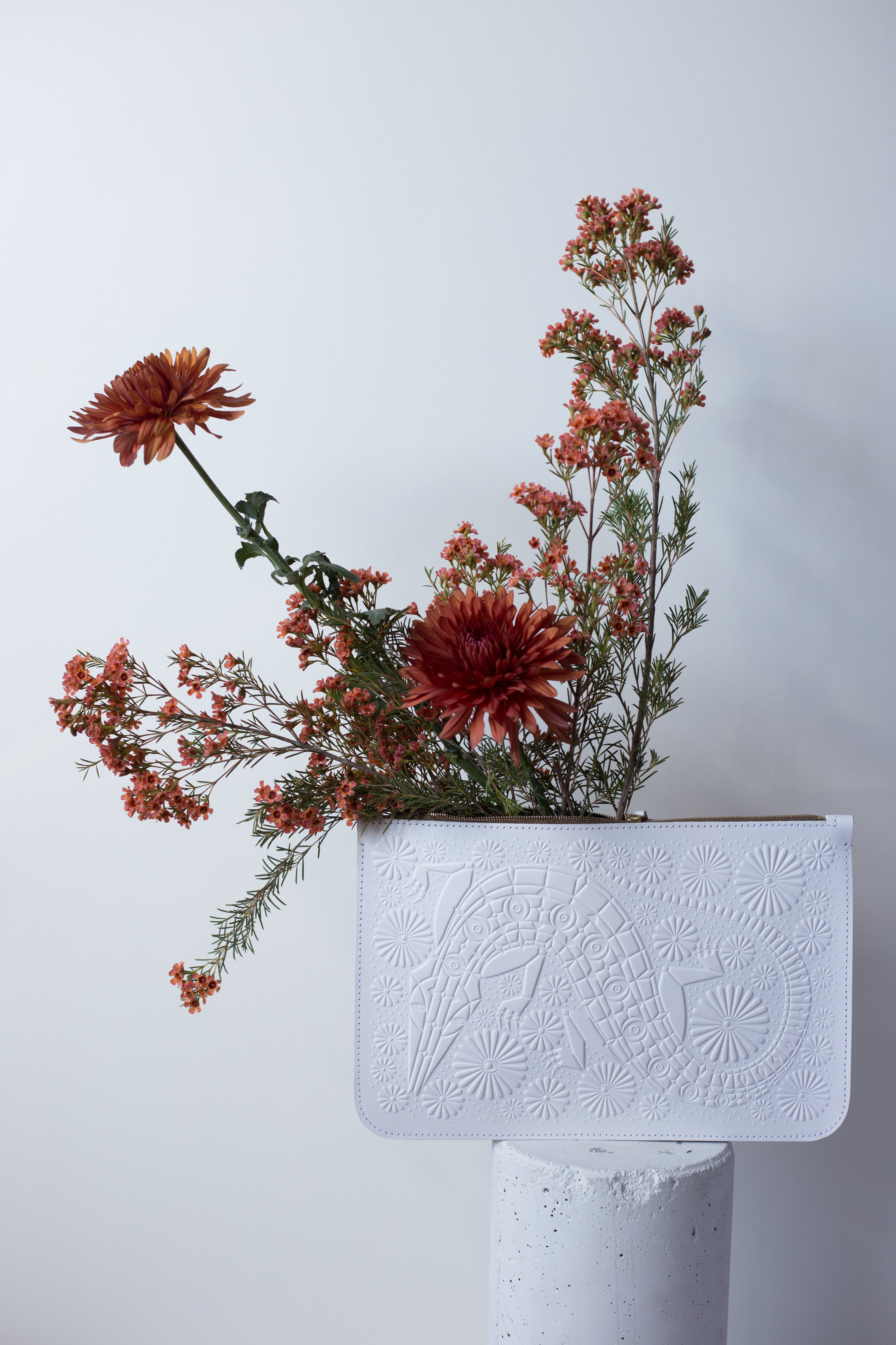 A large white leather clutch with a bold design on it, which includes a large slithering snake or reptile. The clutch is sitting on a white pedestal and has red flowers coming out of the inside of the pouch. the image is captured on a white backdrop. 