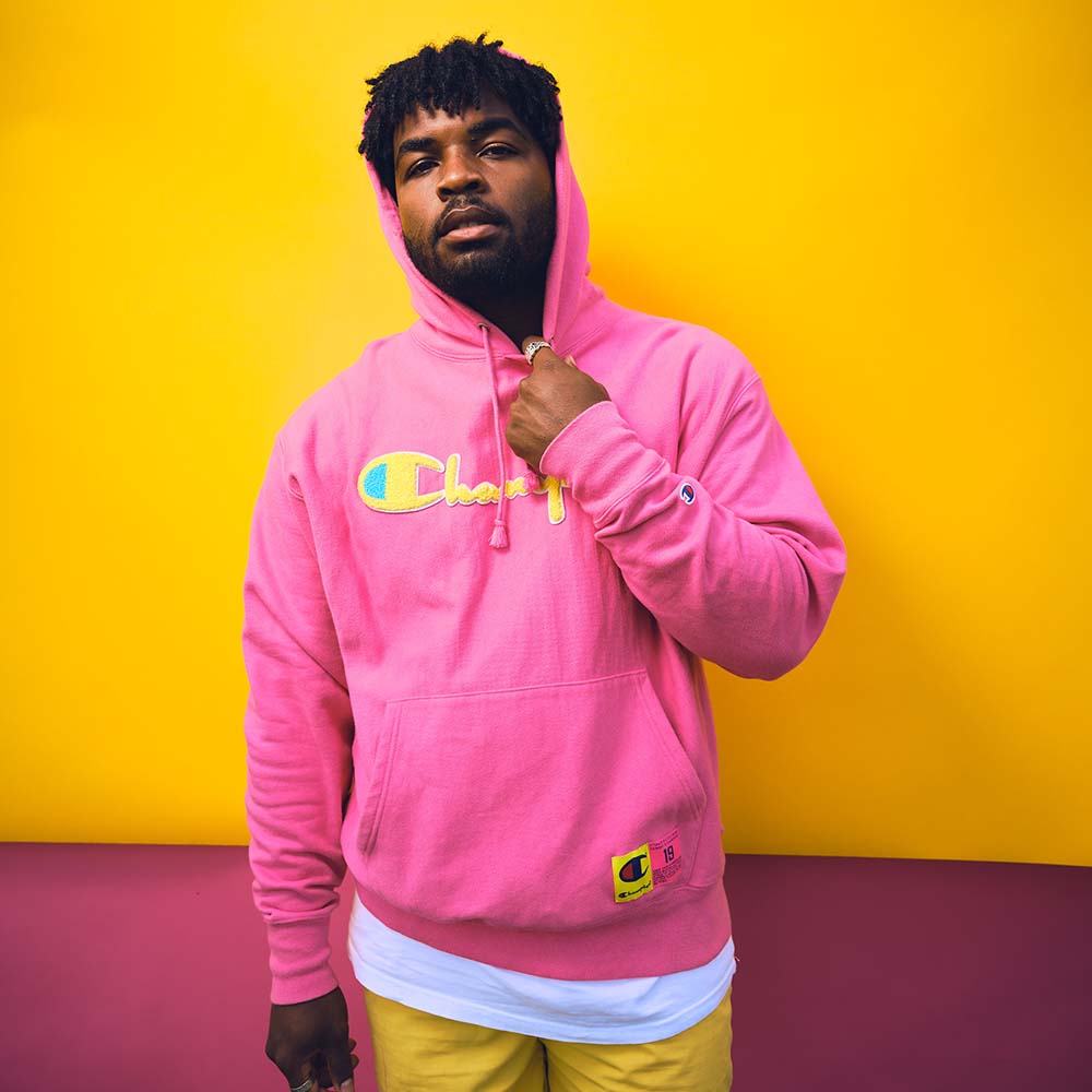 Band photo featuring one person sporting a bright pink hoodie and posing in front of a yellow background.