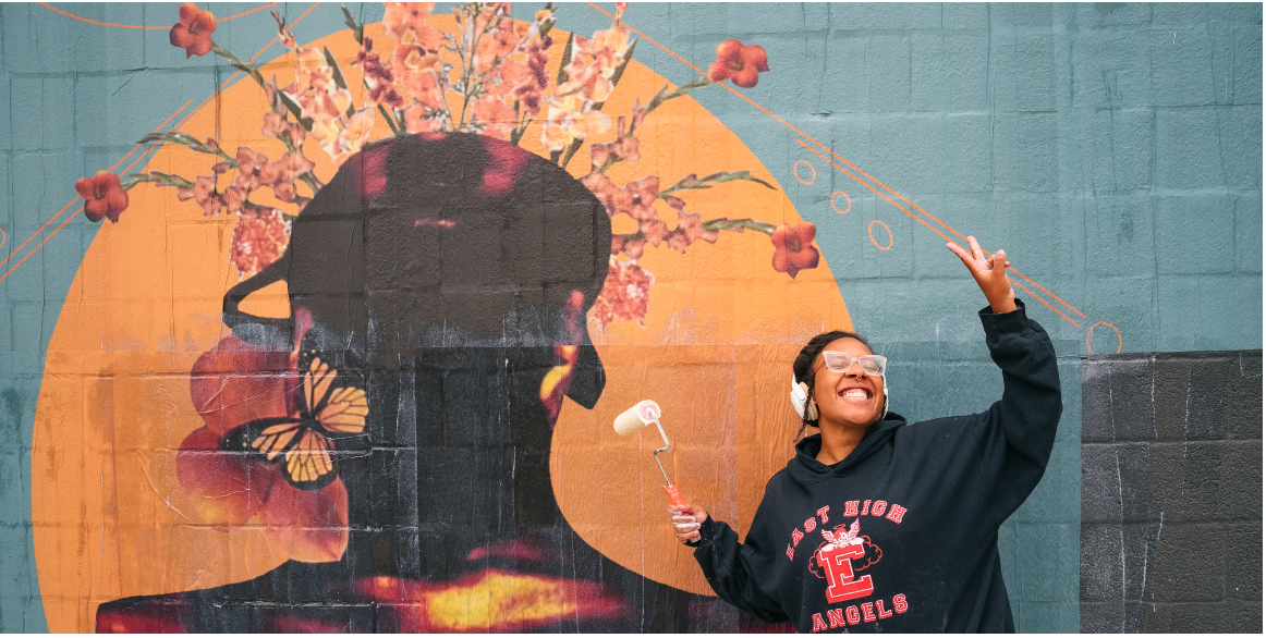 Photo of the artist, Yazzi, with a wide beautiful smile on their face holding a paint brush, wearing glasses and their eyes are closed while throwing up a peace sign. Yazzi is standing in front of their mural at 