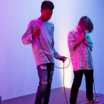 Two young men hold microphones and are lit by pink and blue lighting.