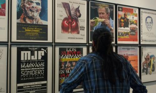 A man looks at framed artworks. He has long black hair and wears a blue flannel shirt. 