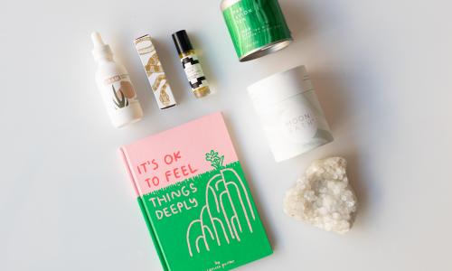 A hotpink and bright green book reads It’s ok to feel things deeply; A white crystal rock; a vial of cbd drops from Dram. Green tea, jasmine, and sugar jems mix from Par Avion Tea; a white container reading moon bath; a scent roller from rootfoot. 