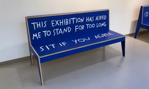A bright blue bench with white, hand-lettered text that reads, "This exhibition has asked me to stand for too long. Sit if you agree"