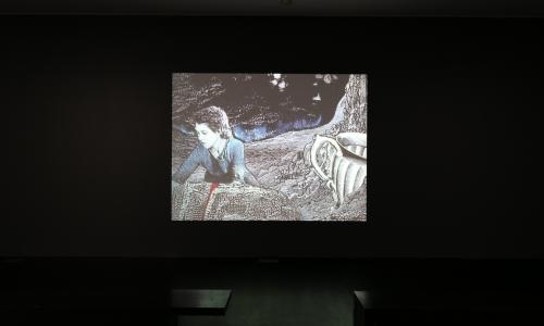 A stop motion film is projected onto a gallery wall. In it, a woman is looking through the ground in a rocky environment. It is half drawn with superimposed paper cutouts. 