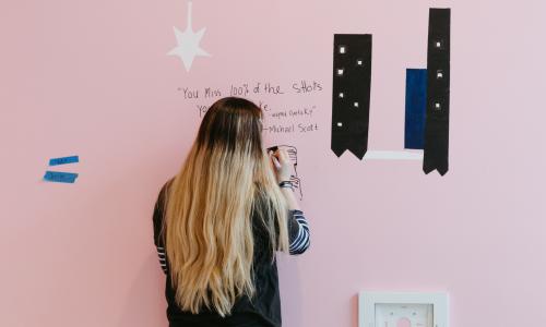 A woman with ombre hair draws an image of a man on a pink wall. The text written in sharpie reads “You miss 100% of the shots you don’t take. -wayne Gretzky” -Michael Scott