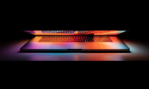 A laptop sits on a surface in a dark room cracked open. The light from the screen emanates onto the desk creating a soft and colorful glow.