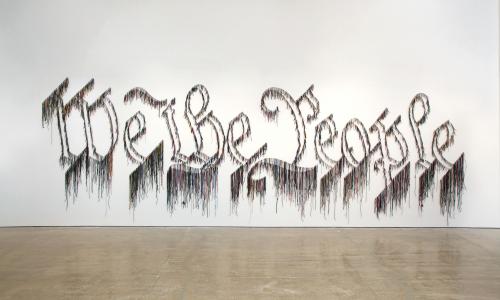 The words “We the People” are spelt with string and attached to a white gallery wall. 
