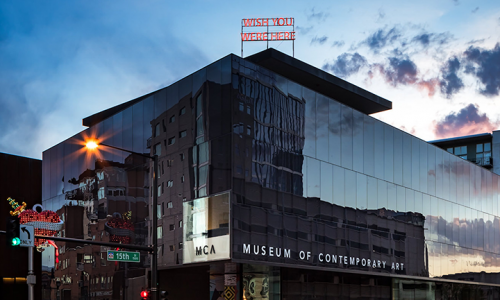 Exterior shot of the Museum of Contemporary Art Denver at sundown. A neon sign in red is displayed on the buildings roof and reads "WISH YOU WERE HERE." The sunset is reflected on the buildings side.