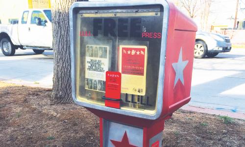 A red newspaper vending machine repurposed into a little library in an outdoor space. It reads LLiLL FREE PRESS. Two pamphlets are visible. “WAR ON PATRIARCHY, WAR ON THE DEATH TECHNOLOGY,” reads the first. The other reads “LEFTIST LEAFLETS IN LITTLE LIBRARIES.”