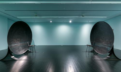 Image of a gallery where two large circular metal sculptures face each other about 10 feet apart. The room is bathed in a teal light and the lights reflect off the dark floor.