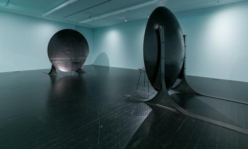 [Image description: An image of an art installation in a white gallery space. The work consists of two chairs, each sitting in front of their own large discs, which are meant to replicate the effects of an acoustic mirror. The discs reflect and focus sound waves in a way that visitors can sit far apart but will hear each other as if they are standing right next to each other.]