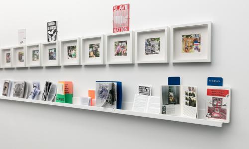 An image of a white wall in one of the MCA Denver galleries. Affixed to the wall are a series of white square frames: inside the frames are photographs of little neighborhood libraries. Underneath is a shelf with colorful zines with different images and graphics on them, with titles ranging from ’SLAVE NATION’ to ‘THE ART AND SCIENCE OF BILLBOARD IMPROVEMENT’.
