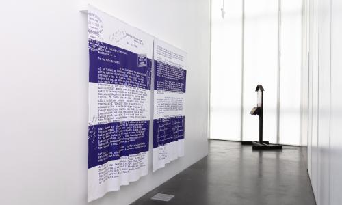 Artist Alan Michelson's, artwork of a reproduction of the December 30, 1924 letter to Calvin Coolidge by the chiefs of the Onondaga Nation, protesting the Indian Citizenship Act and reaffirming Onondaga sovereignty. The reproduction is on a pair of blankets in the colors white and purple and hangs on a white gallery wall with a dark concrete floor.