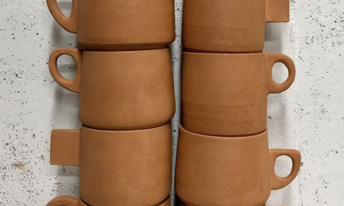 Two stacks of terra-cotta colored mugs waiting to be glazed. Each stack includes five mugs, and each mug has a different style handles on them. The mugs are sitting in front of a speckled black and white backdrop. 