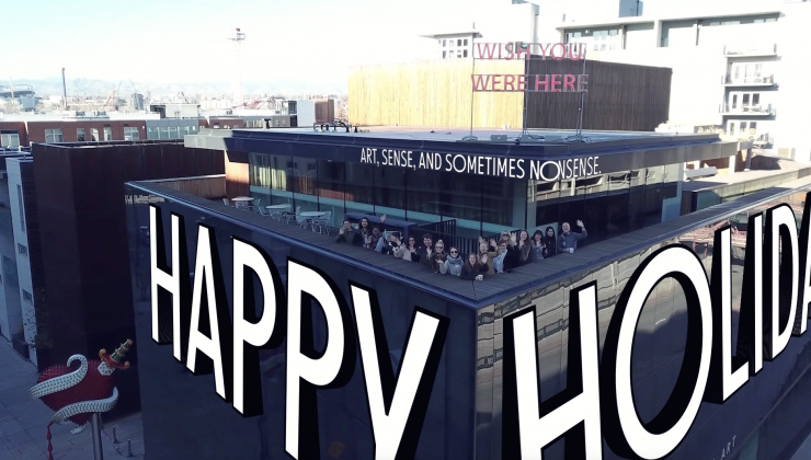 Screenshot of a video. Exterior of MCA Denver taken from a drown shot. Text is overlayed reading "Art sense and sometimes nonsense. Happy Holidays! Museum staff are gathered at the corner of the rooftop. 