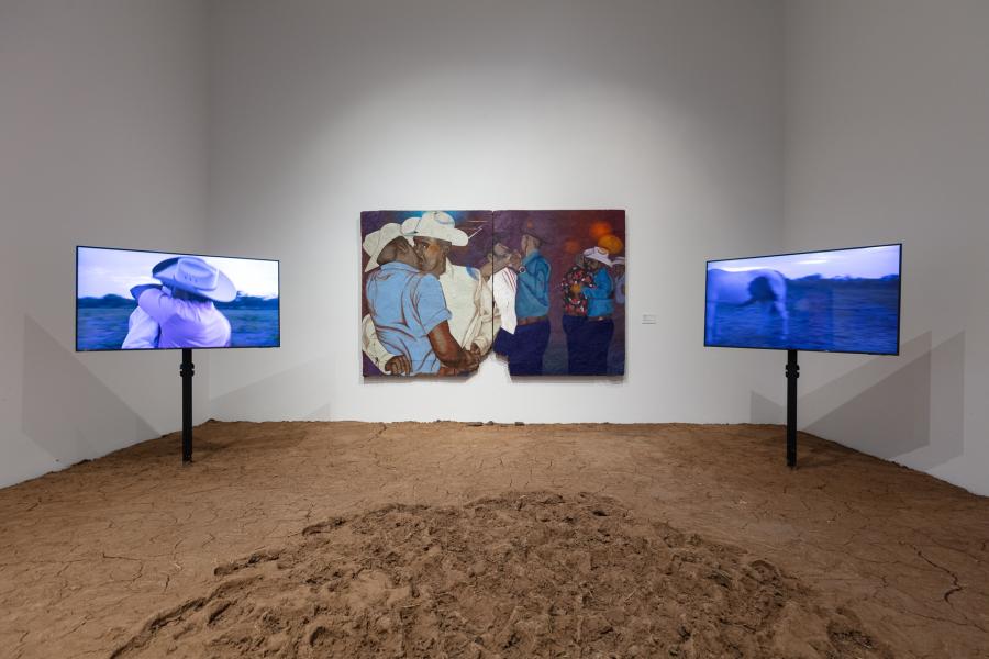 Art installation featuring dirt on the ground, two televisions mounted in the dirt, and a large painting of gay cowboys dancing. 