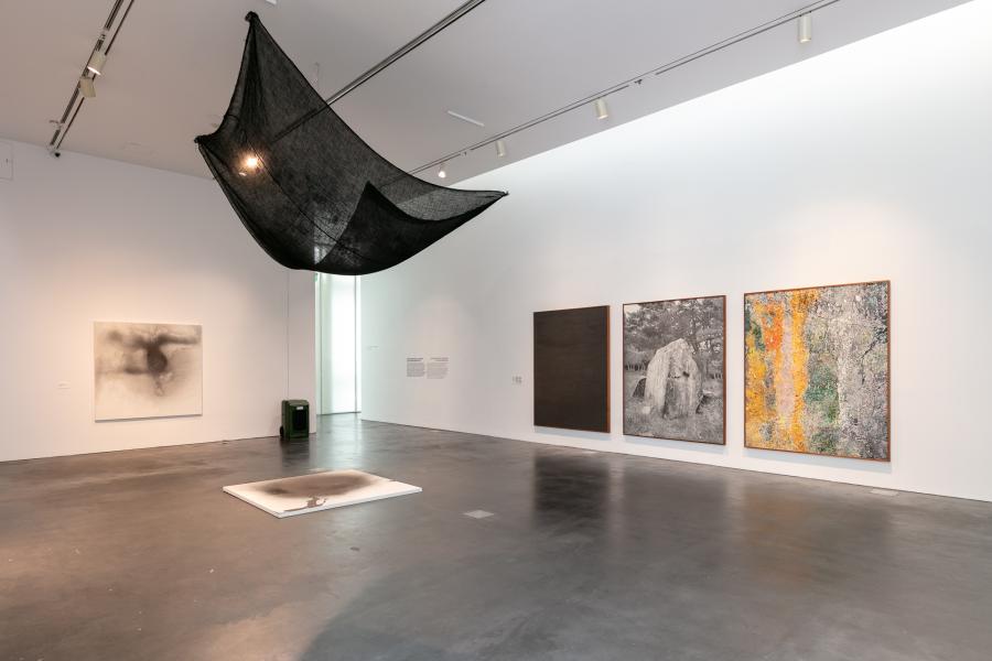 Gallery featuring a large burlap sack hanging from the ceiling. There are three large works of art hung on the wall.