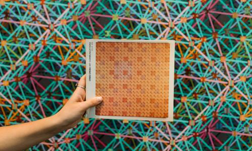 A hand holds a piece of paper with an abstract pattern in tan on it in front of an abstract geometrical artwork.