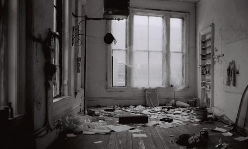 A black and white photo of a messy interior space. There is a window covered in transparent tarp at the far end of the room. Papers and other objects litter the floor. 